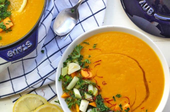 A pot of spicy vegan butternut squash soup in a blue staub cocoette pot on a blue and white checked napkin with saffron and green onions chopped on top of this sunny spicy soup.