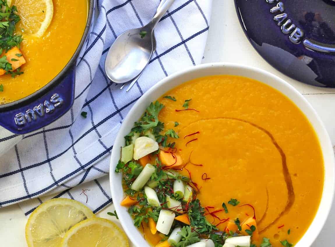 A pot of spicy vegan butternut squash soup in a blue staub cocoette pot on a blue and white checked napkin with saffron and green onions chopped on top of this sunny spicy soup.