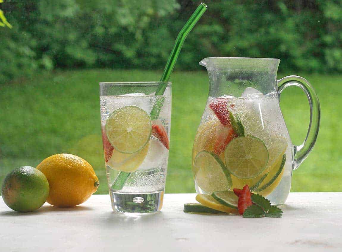 a pitcher and a glass full of infused water and limes and fruits with grass in the background.