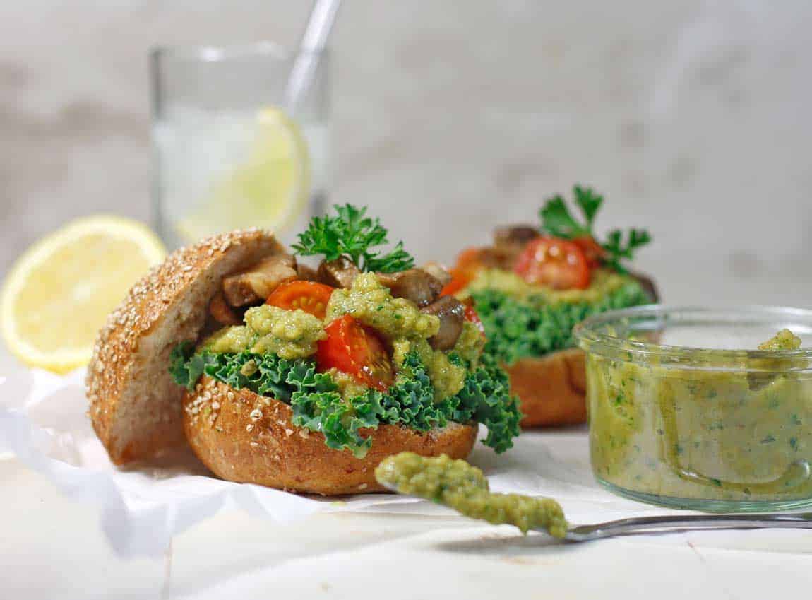 a sandwich stuffed with tomatoes, mushrooms, kale, and spilling over with green olive tapenade which is also on a spoon to the right.