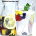 Glass of sparkling water with fruit and veggies floating, a glass straw and a pitcher of sparkling infused water in the background.