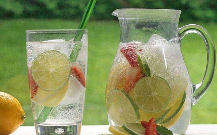 a pitcher of water with infused lime, lemons and strawberries and a full glass with straw