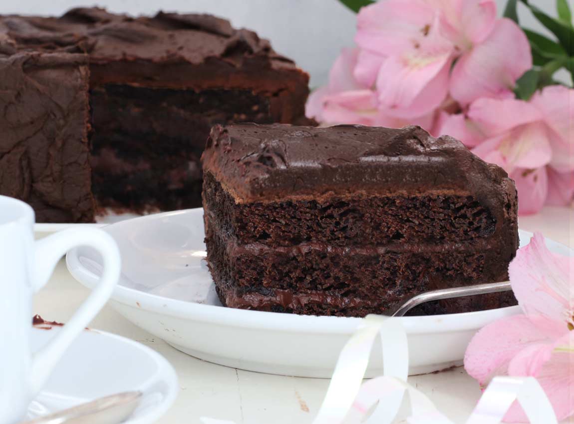 A slice of chocolate cake on a white table with the whole cake behind it and a pink flower behind it.