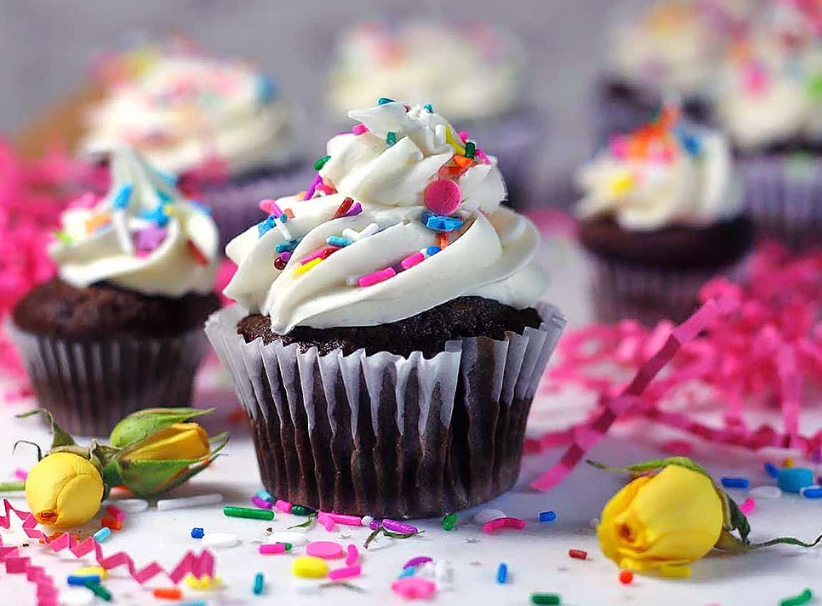 Chocolate cupcake with white icing with rainbow sprinkles on a white table.