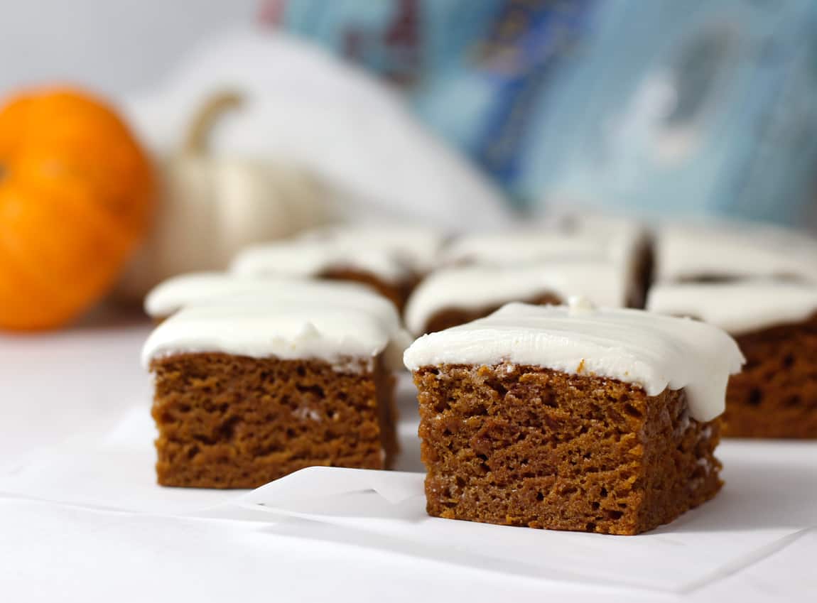 An in-focus slice of healthy pumpkin cake recipe with a vegan cream cheese frosting on top and blurred slices behind it, and an orange pumpkin behind it.