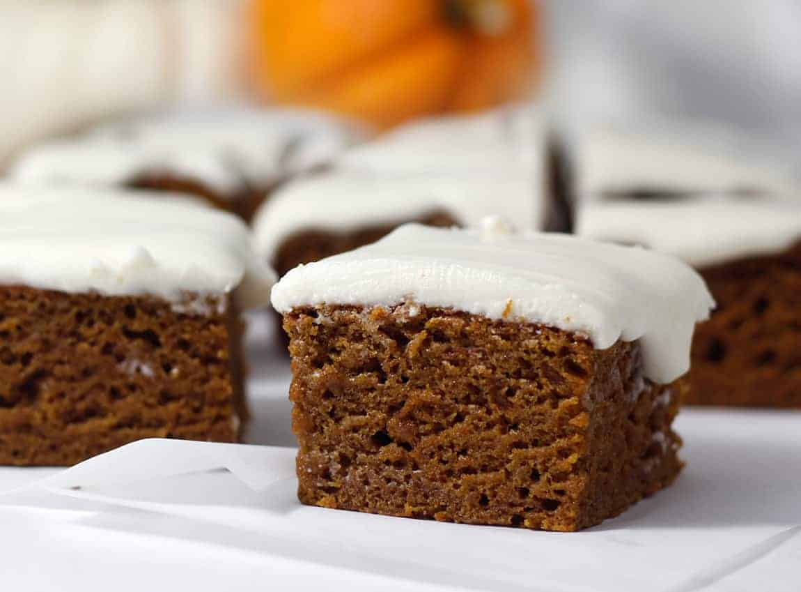 An in-focus slice of healthy pumpkin cake recipe with a vegan cream cheese frosting on top and blurred slices behind it, and an orange pumpkin behind it.