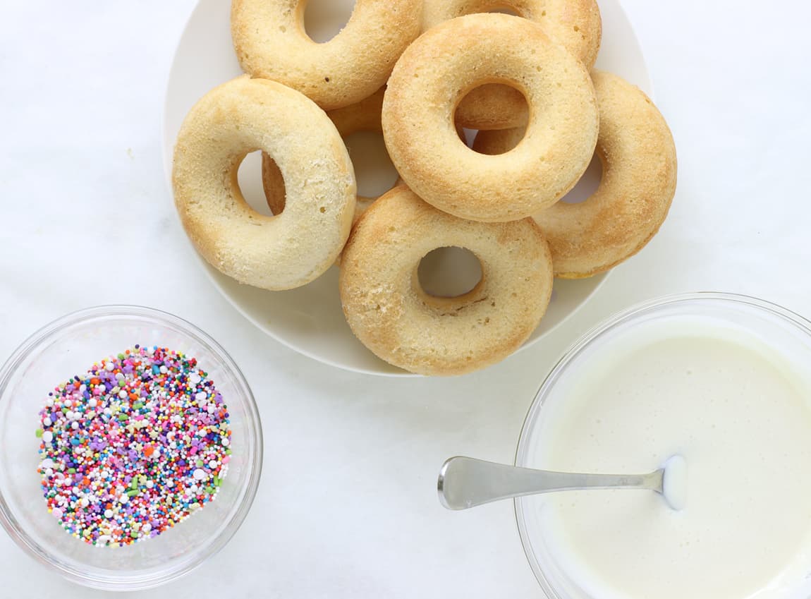 Plain baked donuts with a bowl of icing nearby and a bowl of sprinkles in the corner.