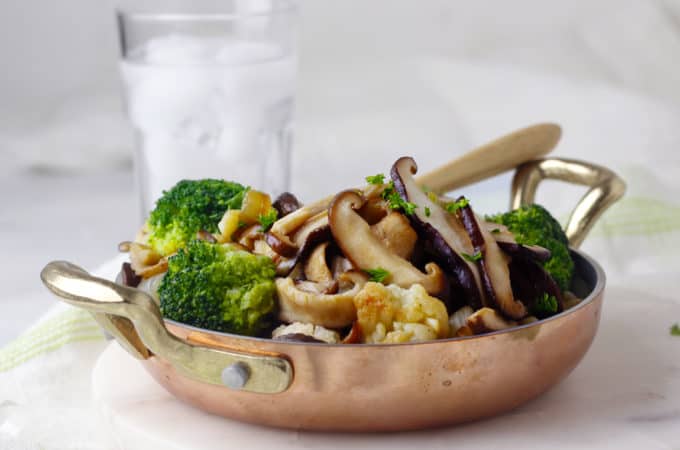 Stir fry mushrooms and broccoli and cauliflower in a brass pan with a glass of water behind it.