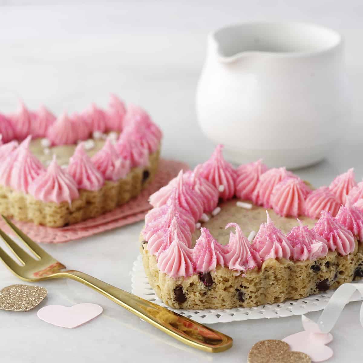 two cookie cake hearts with pink rosette frosting and a white jug behind them