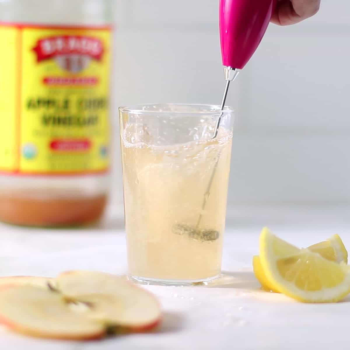 a glass of apple cider vinegar drink with a frother with bottle of acv