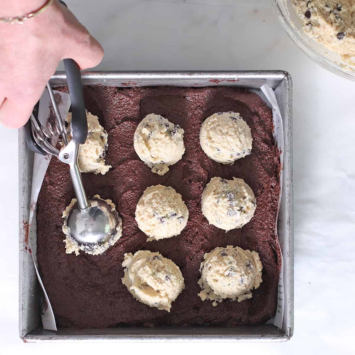 scooping chocolate chip cookie batter onto brownie batter in brookie pan.